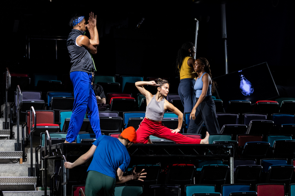 a group of young dancers dressed in bright colored workout clothes dance, balance, touch and walk through the seats of the theater